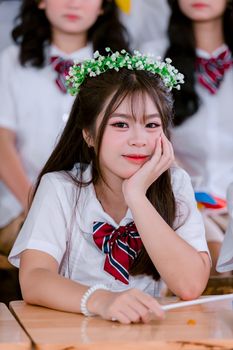 A cheerful Asian schoolgirl wearing a flower crown, smiling in a classroom setting.