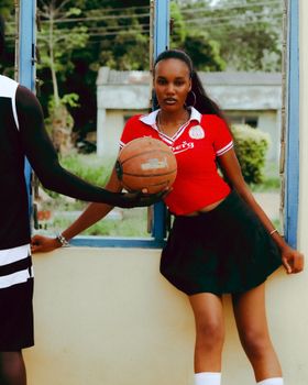 Confident woman poses with basketball by window, wearing red top and black skirt, outdoors.