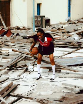 A young woman dribbles a basketball in an urban setting filled with debris, showcasing resilience.