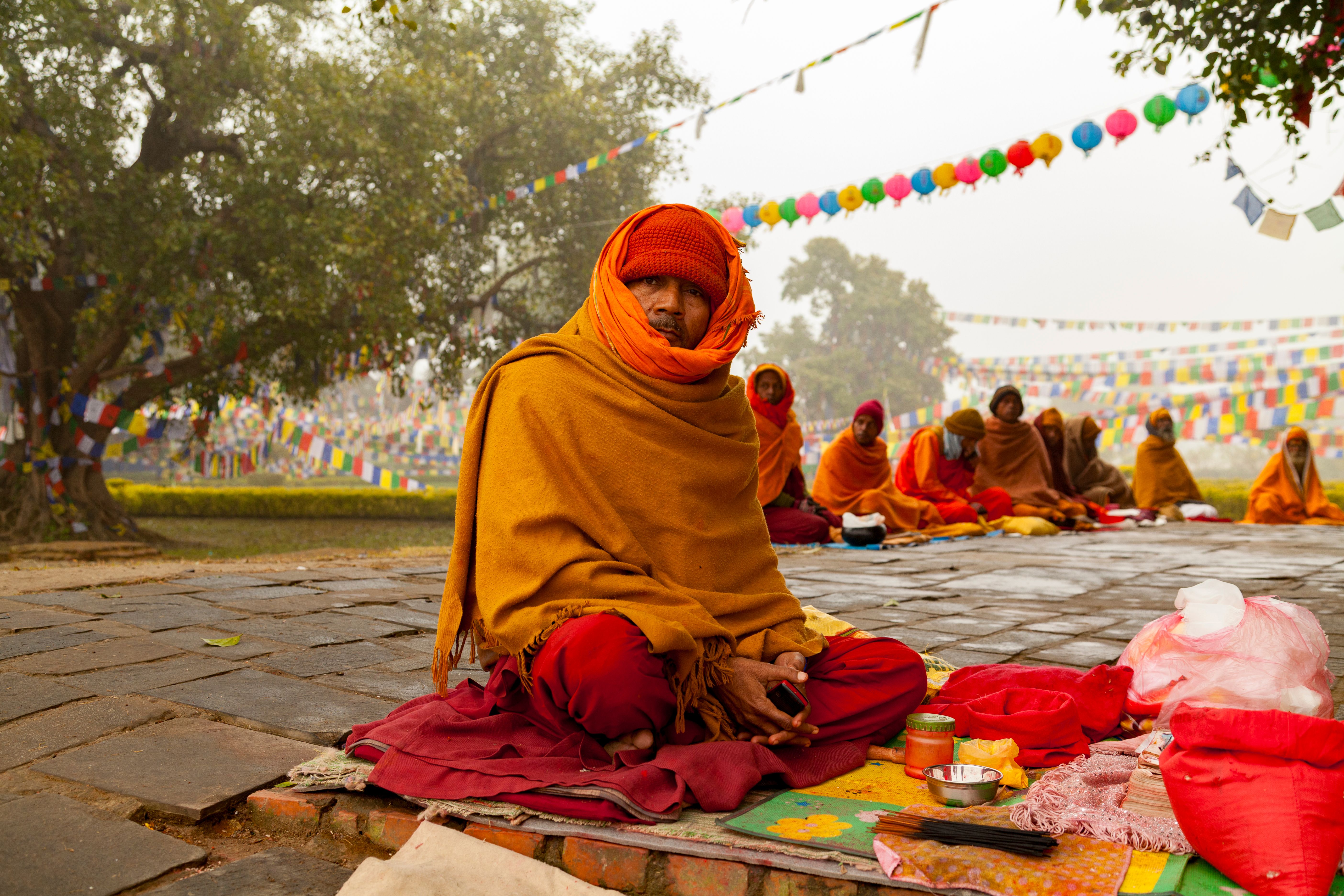 Monks in vibrant robes meditate outdoors in Lumbini, Nepal, surrounded by prayer flags.