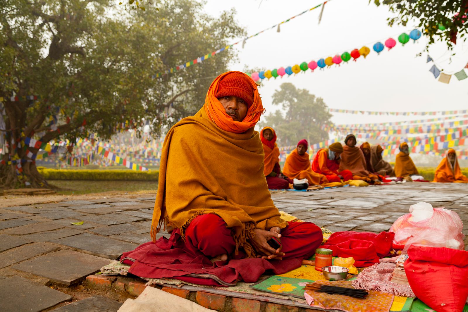 Pilgrims wrapped in saffron shawls seated in a row on stone paving under prayer flags in Lumbini on a misty morning