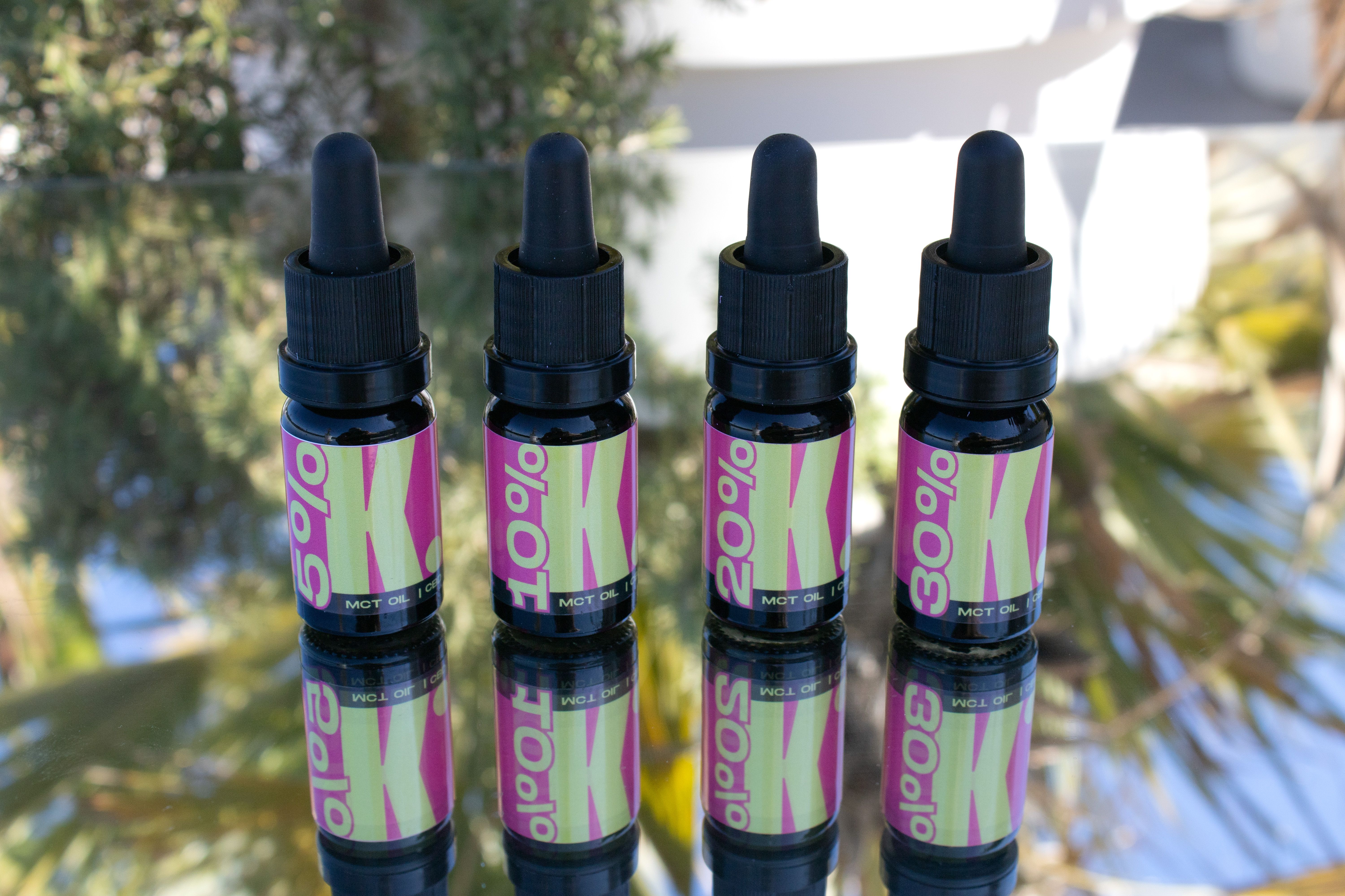 Four CBD oil bottles reflecting on a surface with outdoor palms.