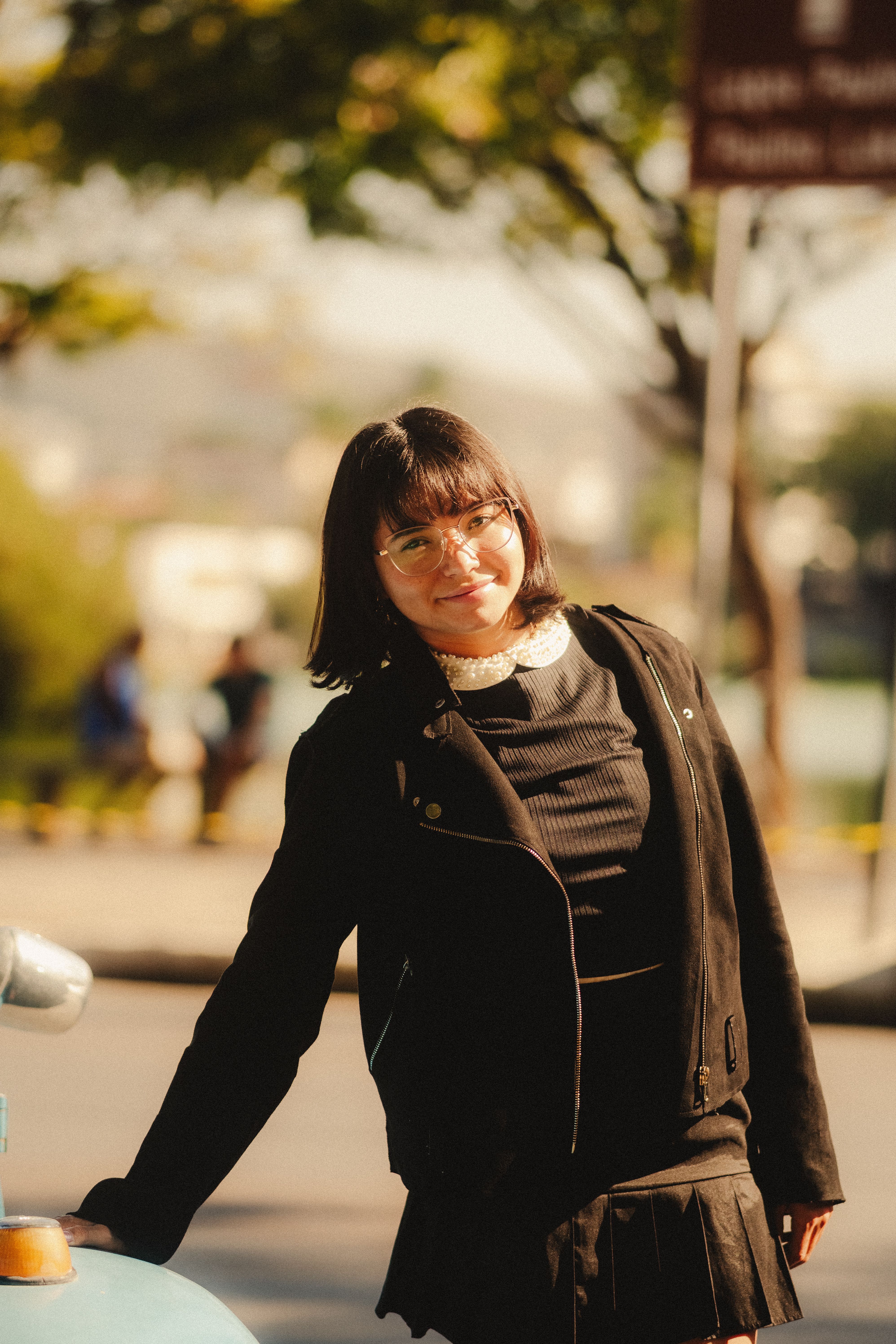 A young woman in a black outfit smiling warmly in an autumn outdoor setting.