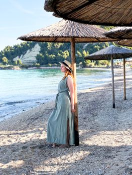A woman in a sun hat and dress enjoys a sunny day on a serene beach with straw umbrellas.