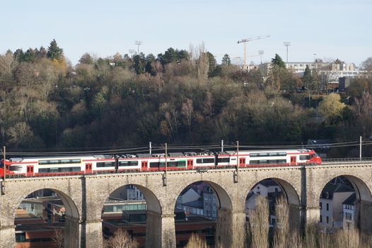 A red train travels over a historic viaduct with a forested backdrop in Luxembourg, showcasing European architecture.