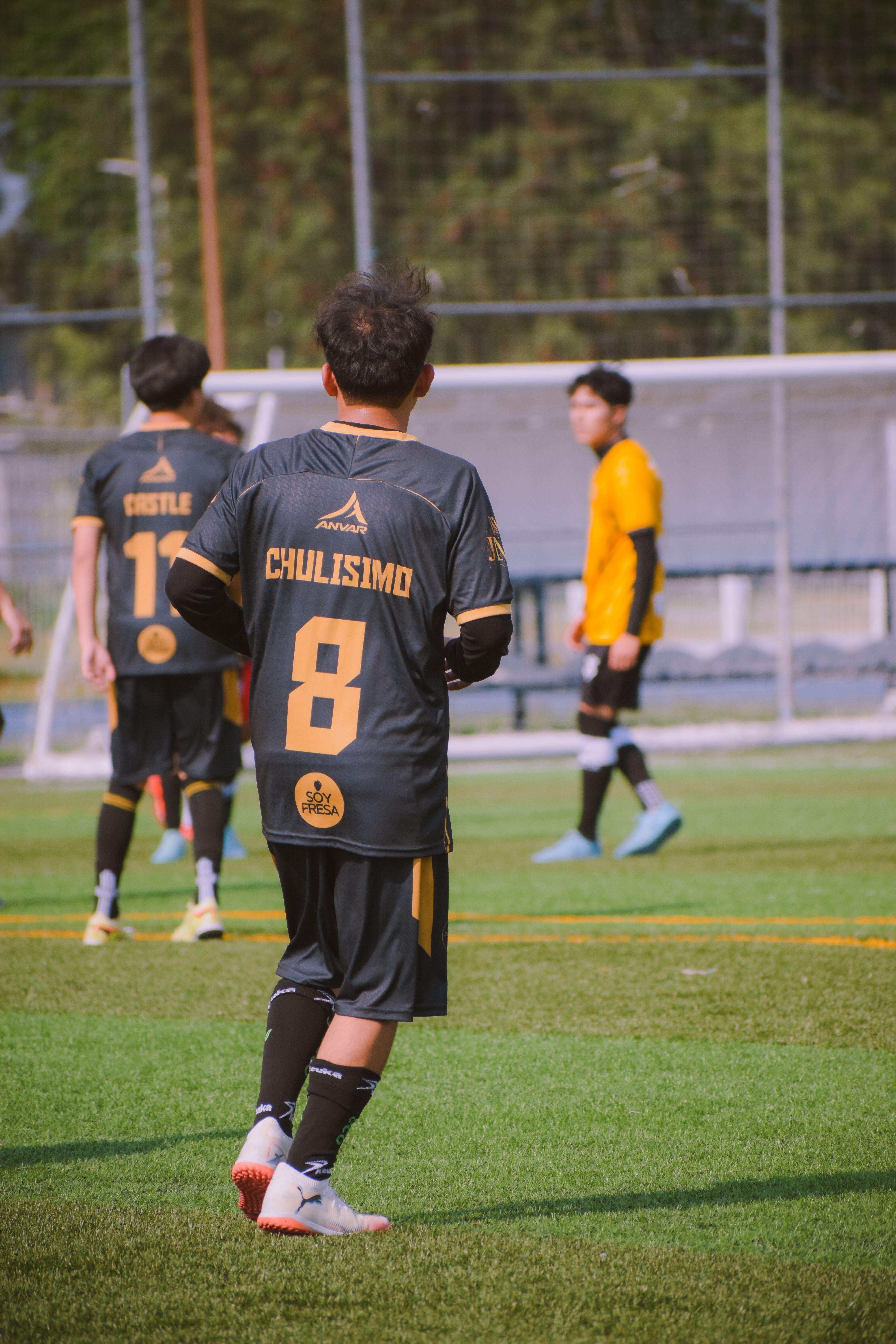 A lively soccer match with players in black and yellow jerseys in action on a sunny day.
