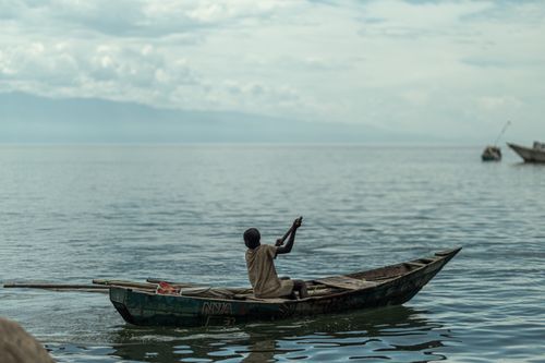 Free A lone fisherman navigates a wooden boat on a serene lake under a cloudy sky. Stock Photo