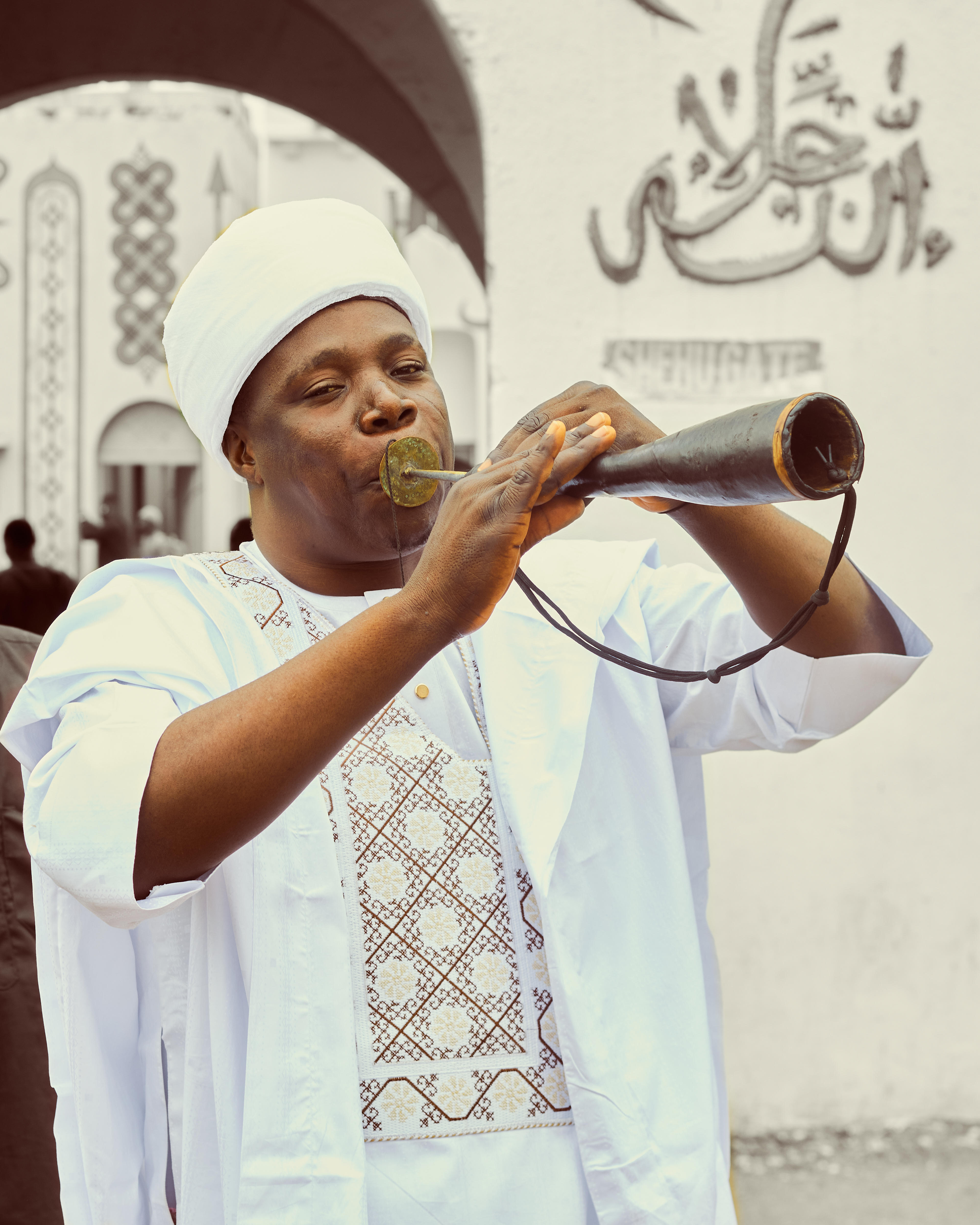 African man in white traditional attire playing a horn in front of an artistic backdrop.