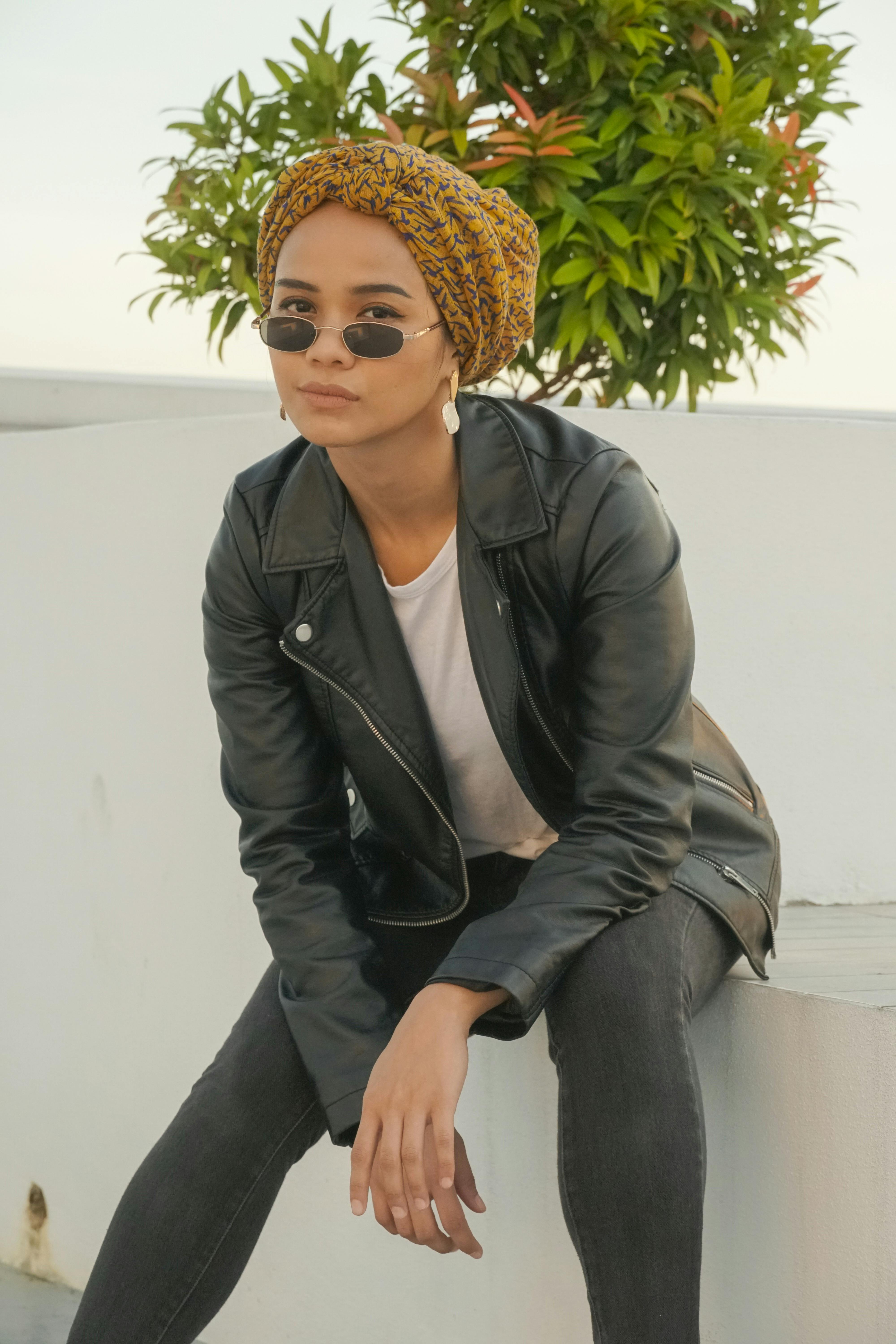 Free A fashionable woman in a leather jacket and headscarf sitting outdoors, exuding modern style and confidence. Stock Photo