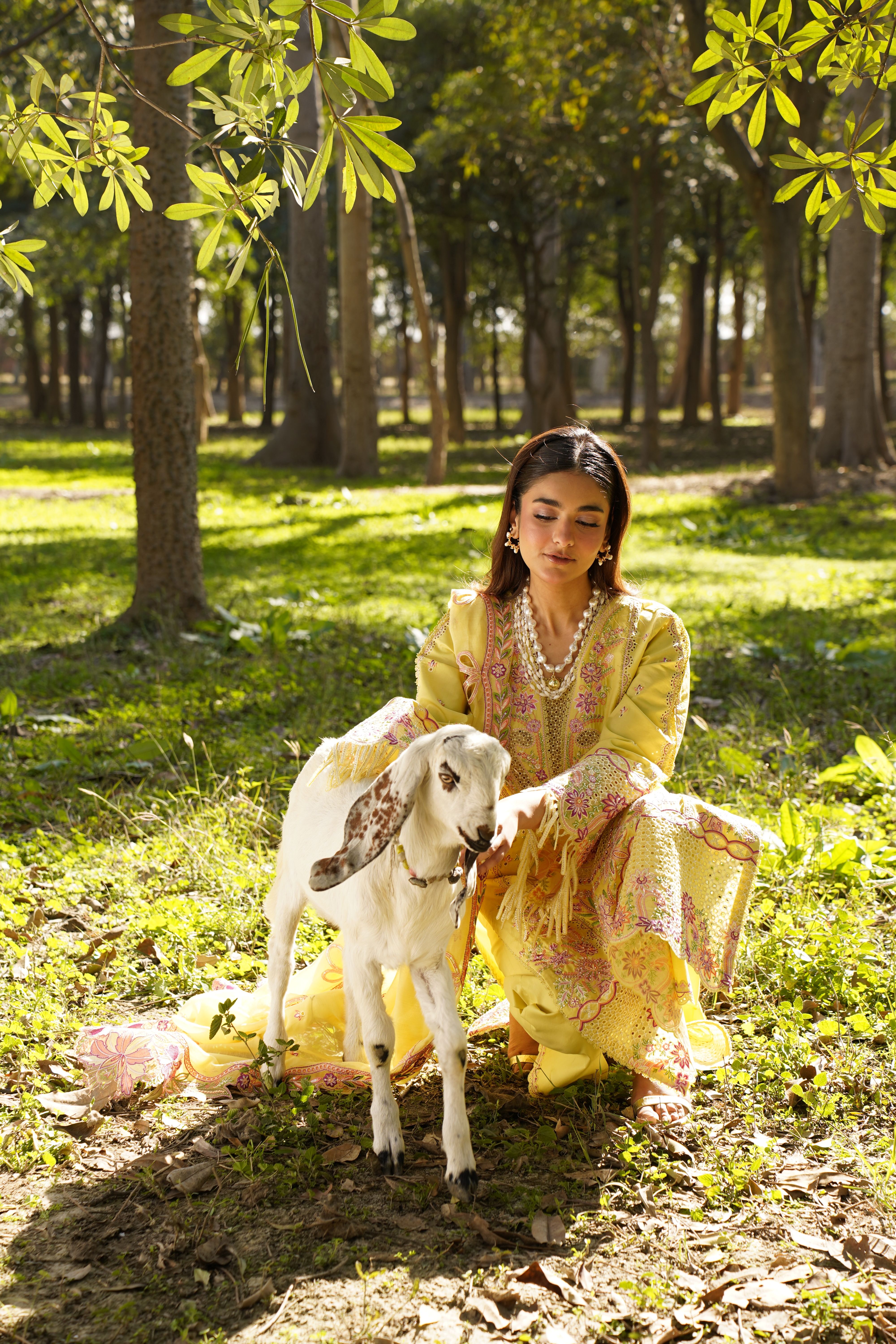 A woman in a traditional dress is sitting with a goat in a sunlit forest setting.