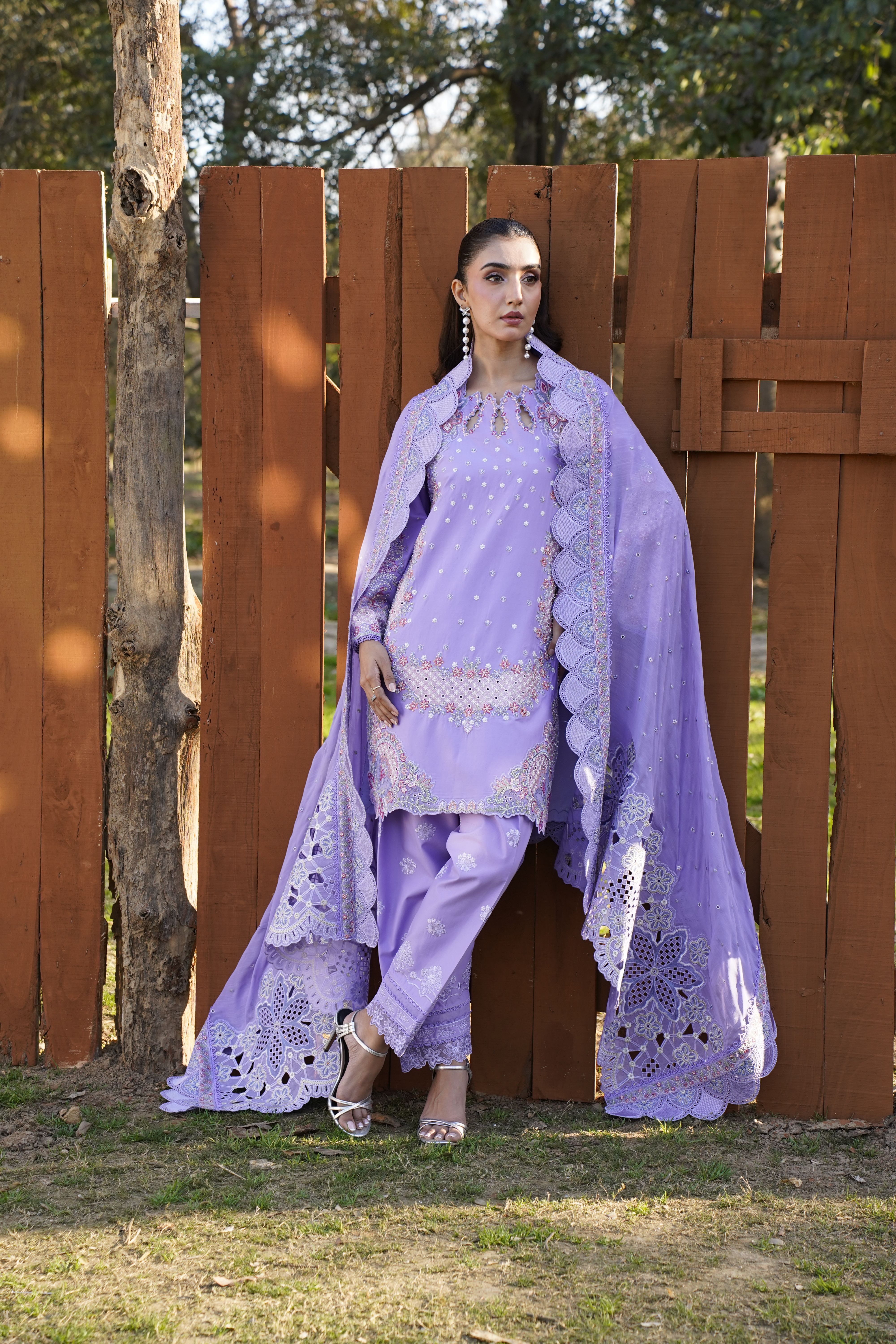 Model in an elegant lavender dress posing outdoors by a wooden fence.
