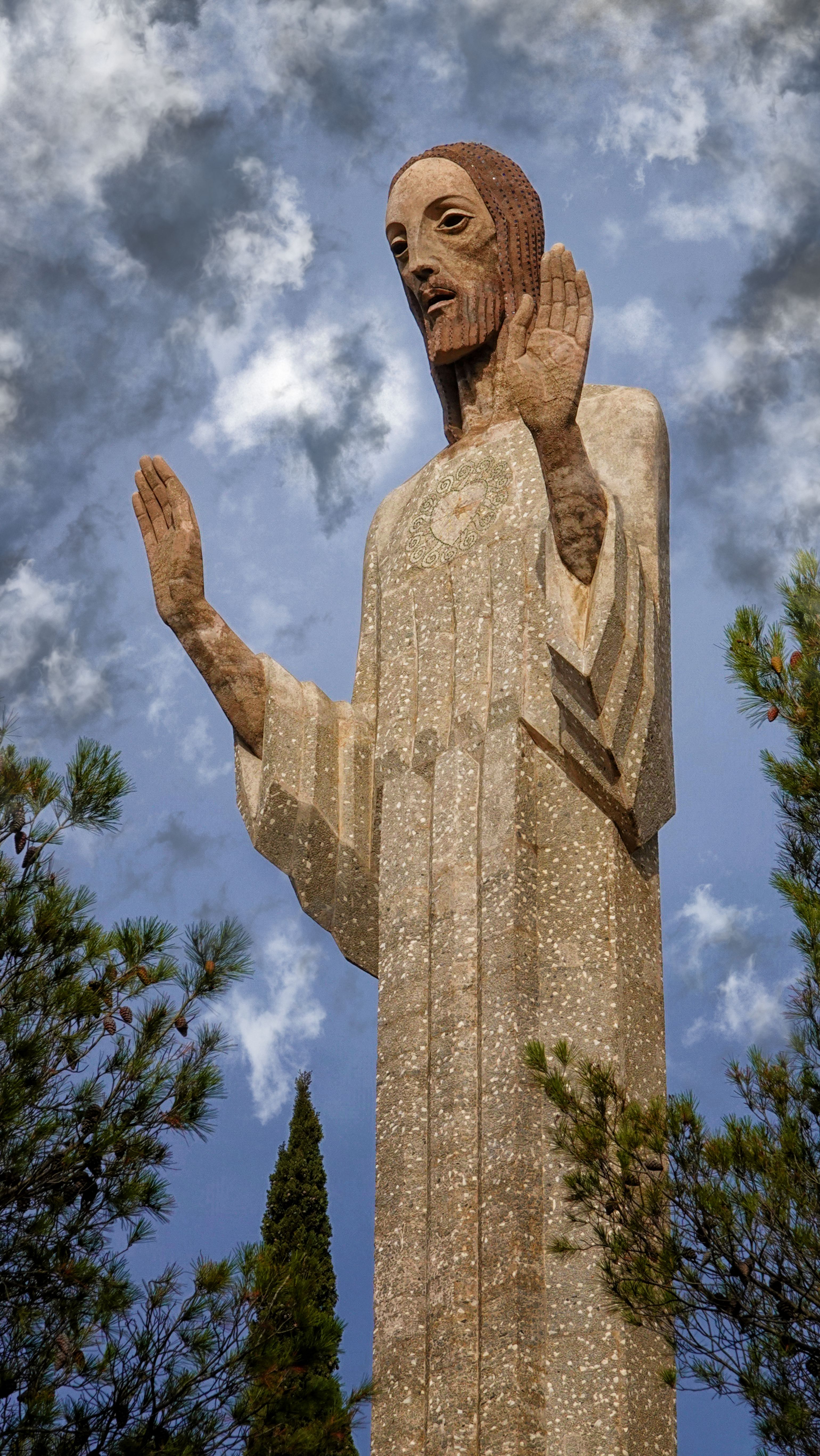 A majestic statue of Jesus Christ stands tall against a dramatic cloudy sky, surrounded by trees.