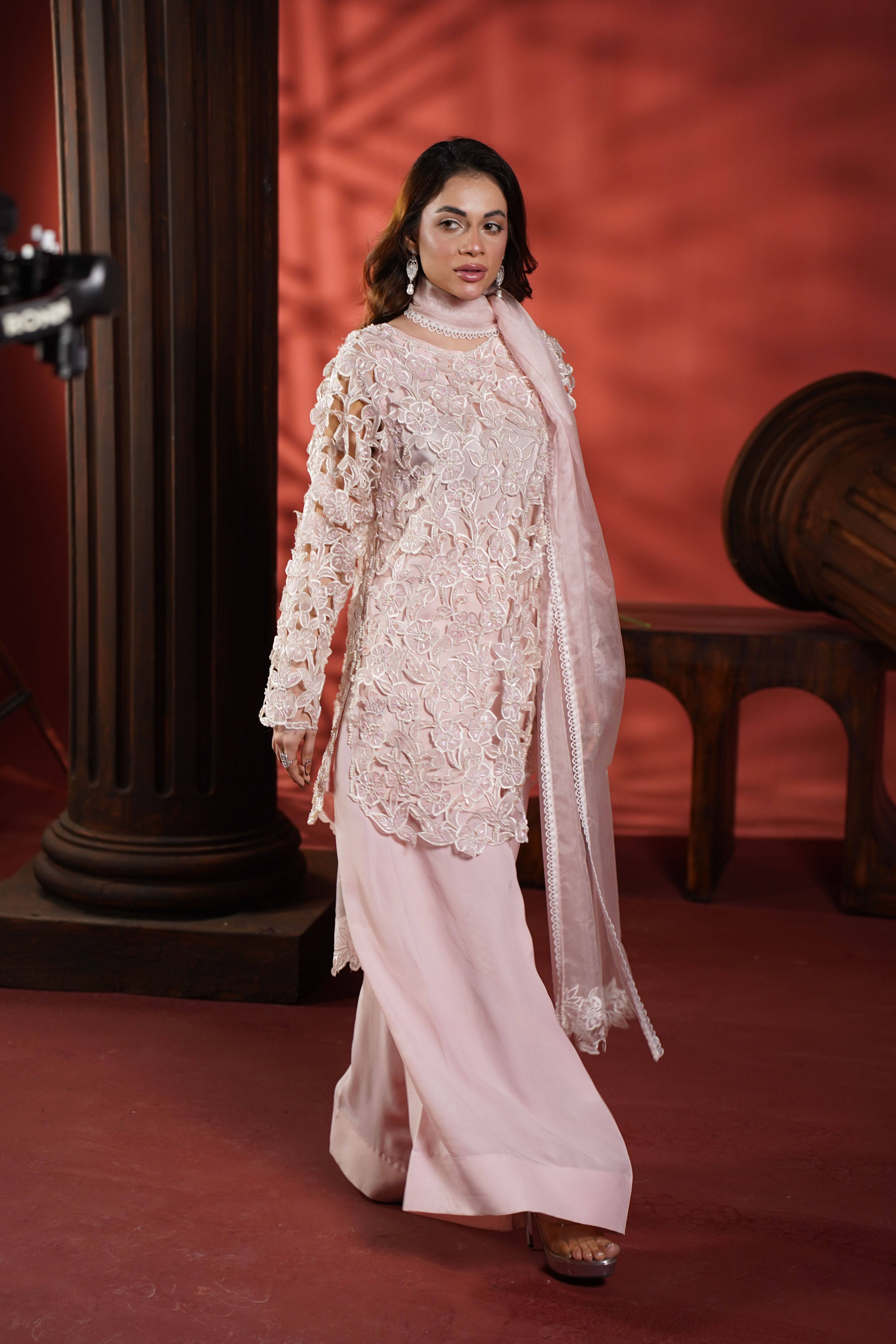Fashion model in ornate soft pink outfit walking past a classic column indoors.