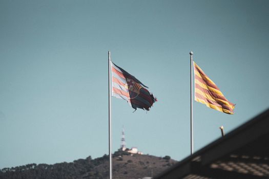 Flags of FC Barcelona and Catalonia waving on poles against a clear sky with distant hills.