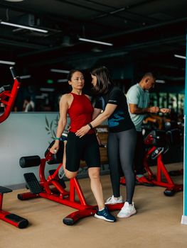 A personal trainer helps a woman with exercise equipment in a modern gym setting.