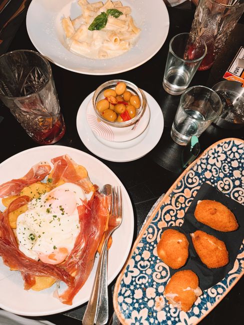 A pile of golden-brown fried Spanish ham croquettes on a white plate, one broken open to show the creamy interior, with a small dish of aioli alongside.