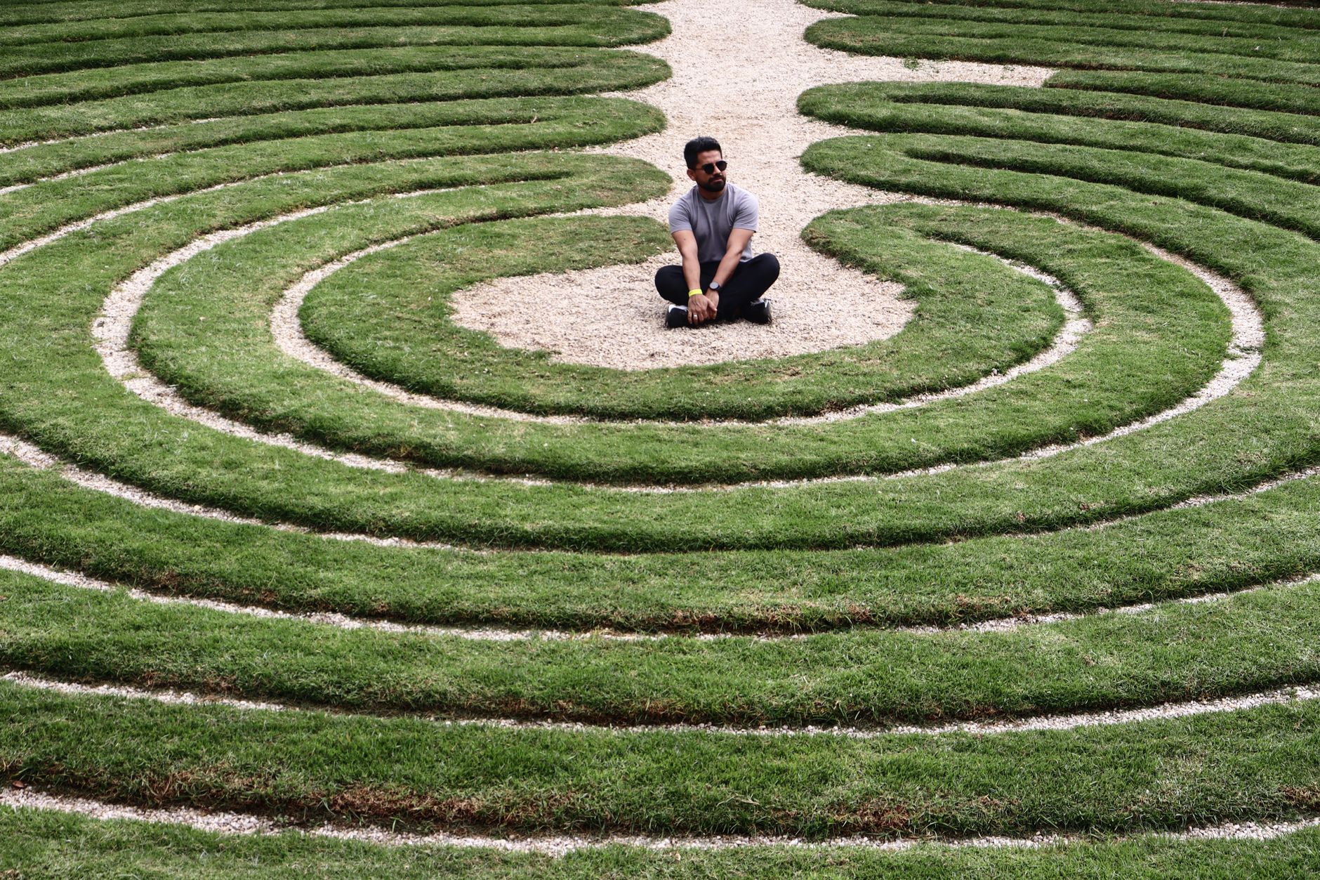A man sits in the center of a spiral grass maze on a sunny day.