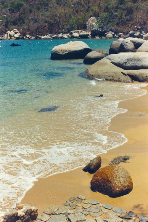 Free A serene beach scene with clear blue water and large shoreline rocks. Stock Photo