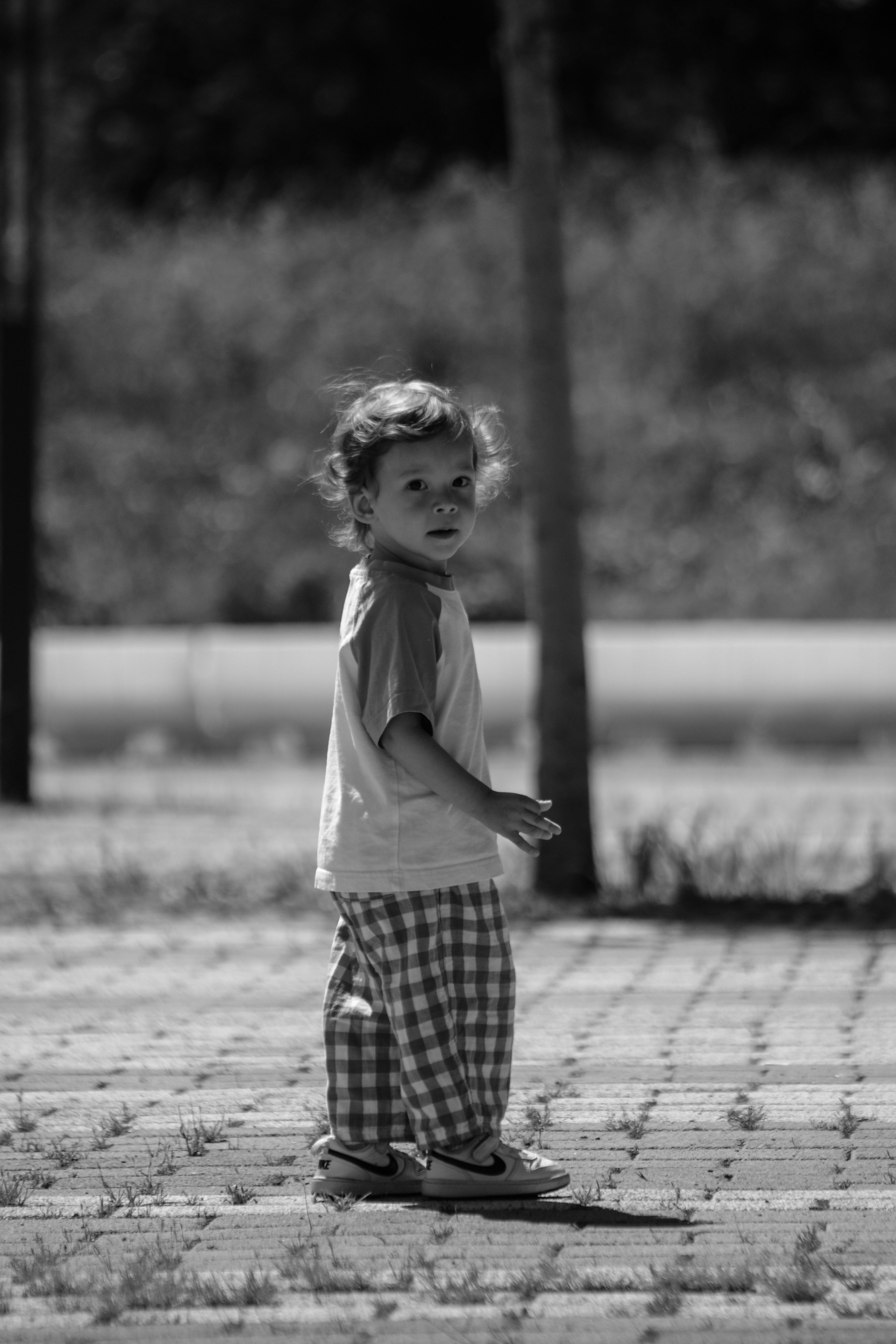 Uma foto espontânea em preto e branco de uma criança pequena em pé em um caminho pavimentado ao ar livre.