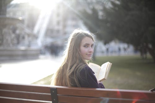 Free Back view of peaceful female in casual jacket sitting on wooden bench in city park and enjoying story while relaxing and looking over shoulder Stock Photo