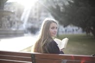 Back view of peaceful female in casual jacket sitting on wooden bench in city park and enjoying story while relaxing and looking over shoulder