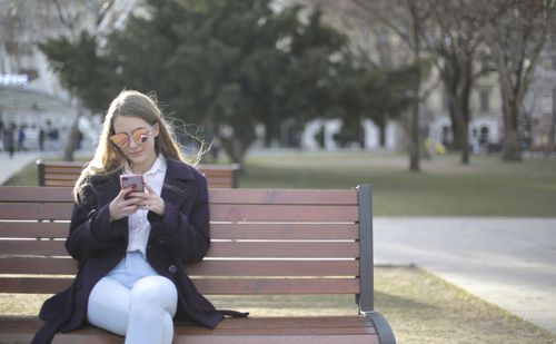 Free Young woman using smartphone while sitting on a park bench wearing sunglasses and a violet coat. Stock Photo