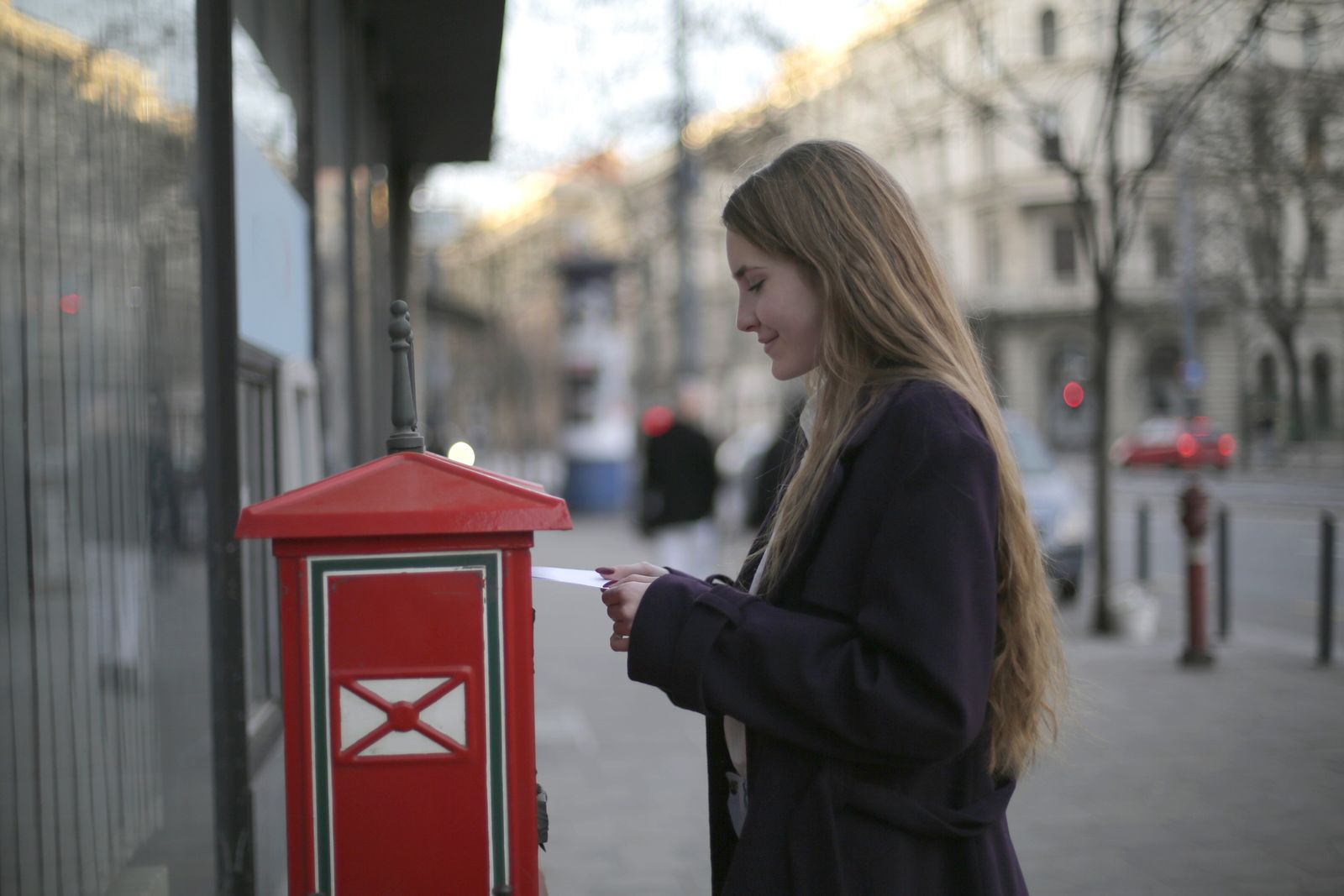 A young woman posting a letter into a red post box on a city street