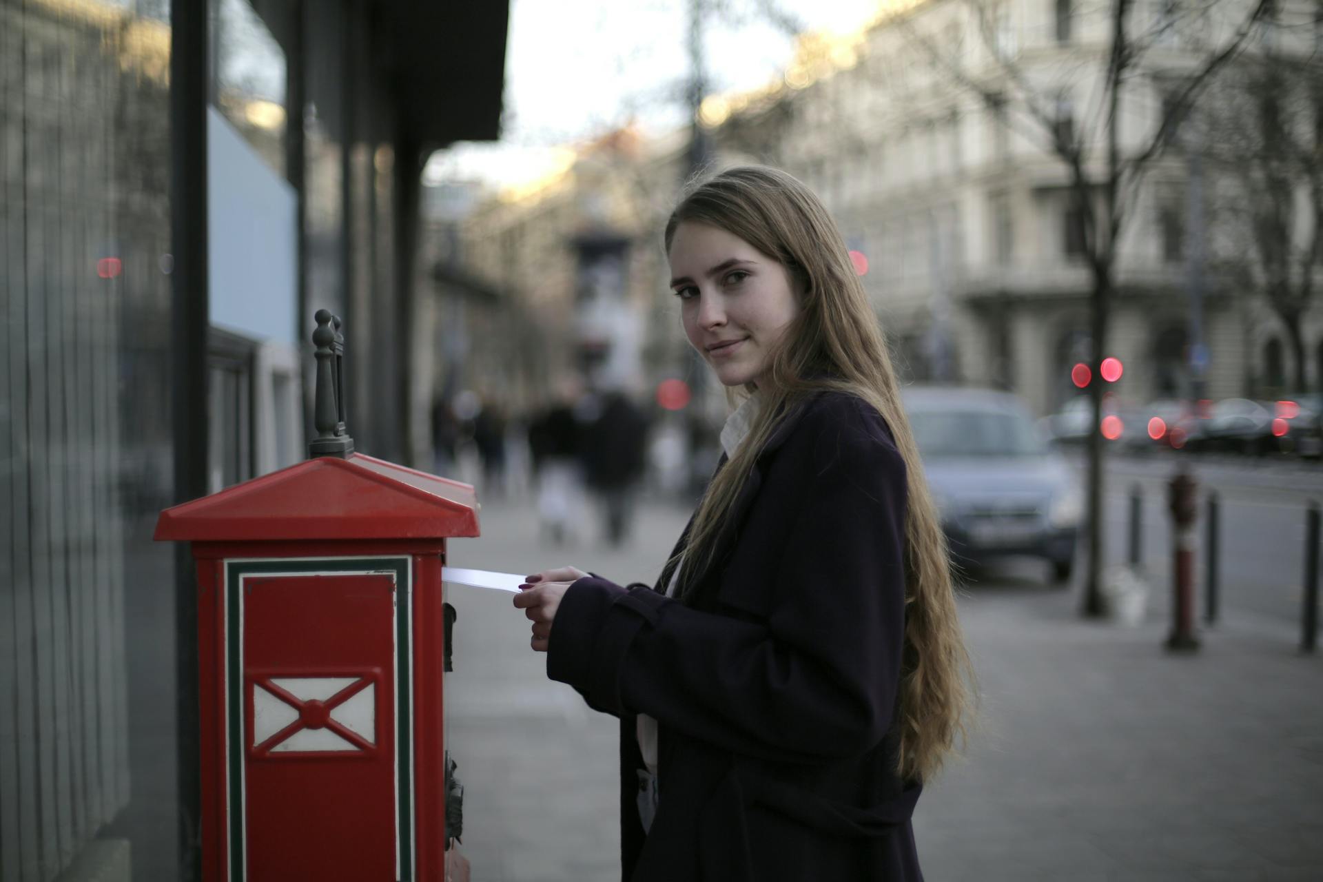 Young woman mailing a letter in a red mailbox on a city street during the day.