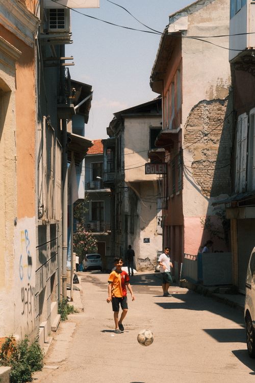 Free Children enjoy a casual game of soccer in a sunlit alley, surrounded by rustic buildings. Stock Photo