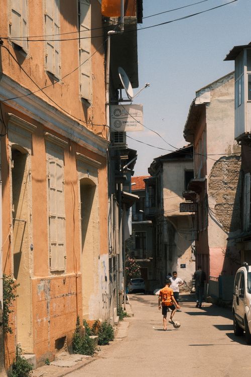 Free Two kids play soccer on a sunny day in a quaint Mediterranean alley, surrounded by rustic buildings. Stock Photo