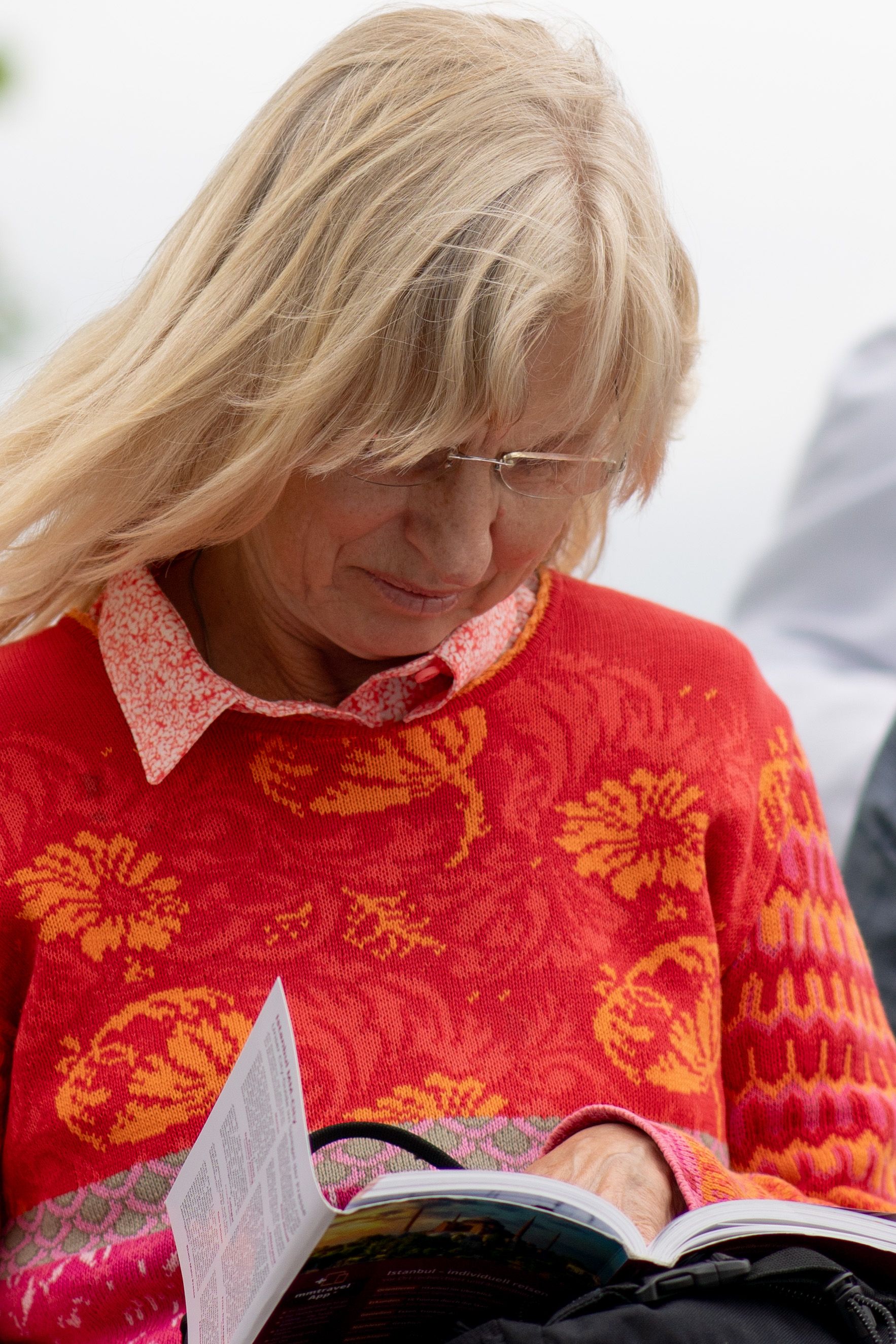 Elderly woman in vibrant sweater peacefully reading outdoors on a cloudy day.
