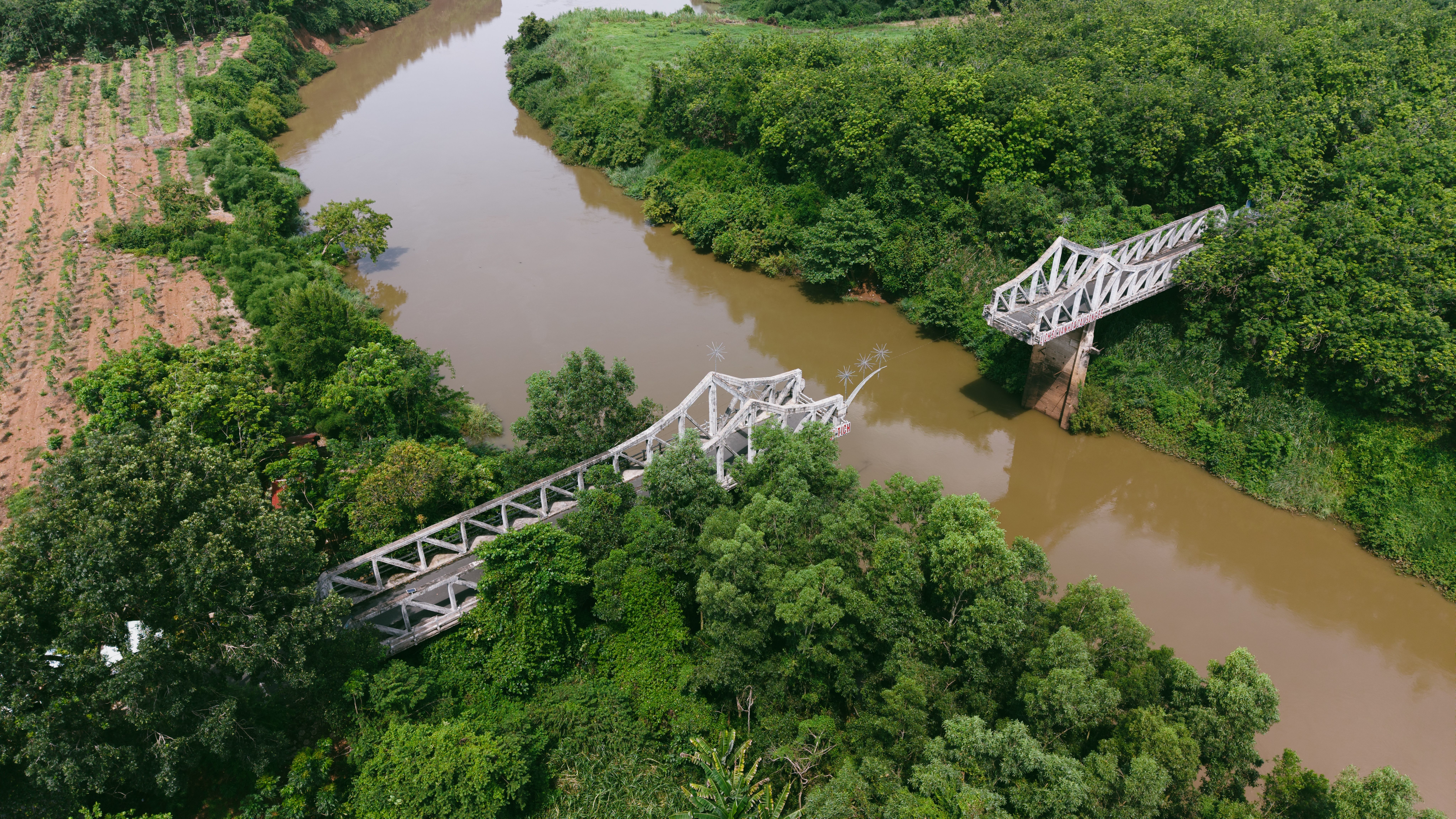 Aerial shot of a broken bridge spanning a river amidst lush greenery and farmland.