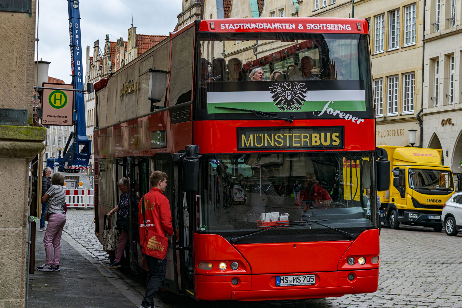 Red double-decker Münster city tour bus at Prinzipalmarkt, bustling with tourists exploring the historic district.