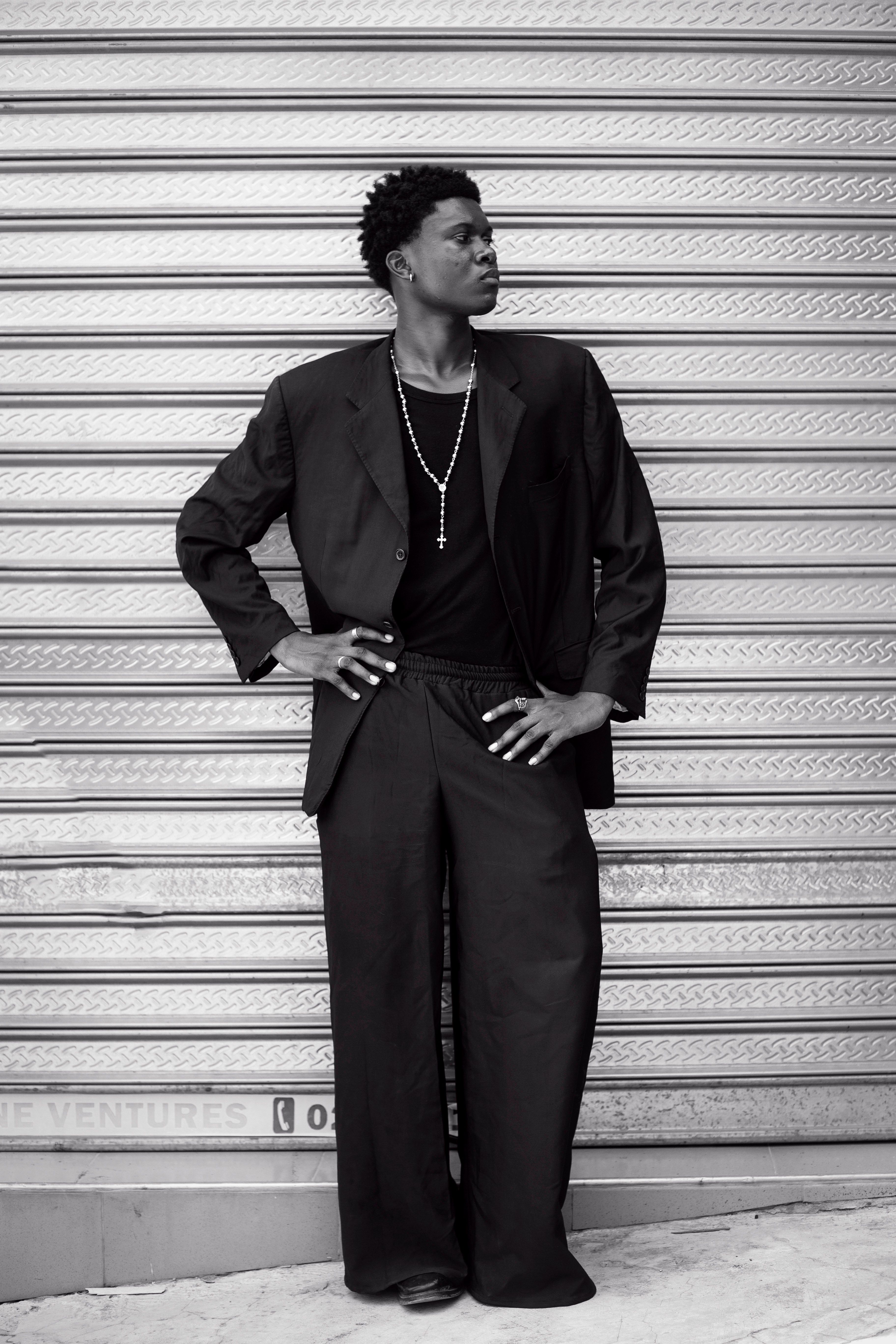 Black and white portrait of a stylish man in a suit standing confidently against a corrugated metal wall.