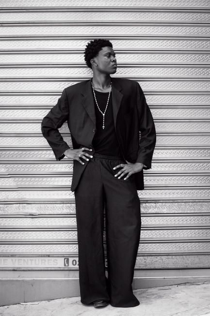 Black and white portrait of a stylish man in a suit standing confidently against a corrugated metal wall.