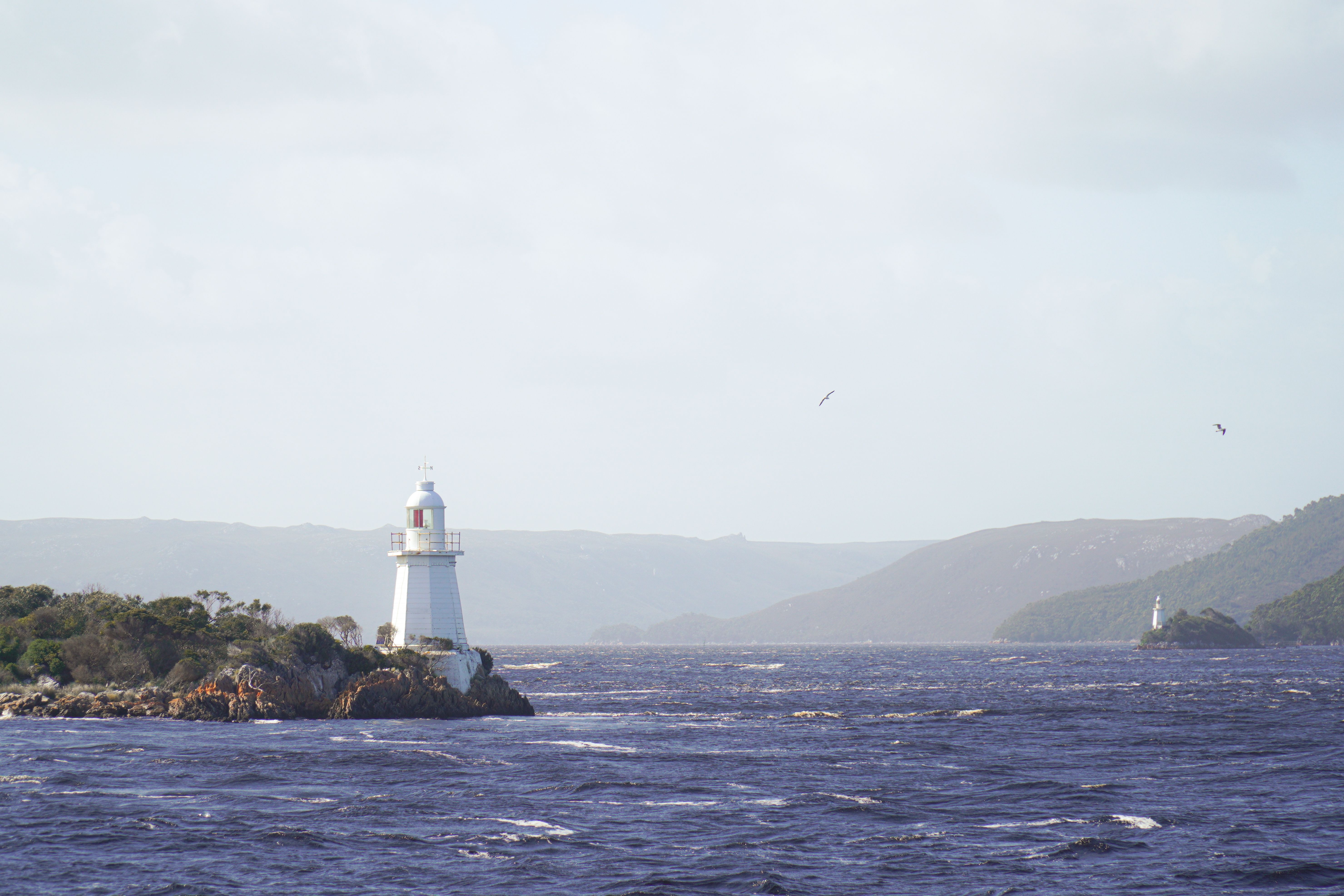 Distant view of lighthouses in Strahan, Tasmania against a backdrop of rugged landscapes and ocean waves.