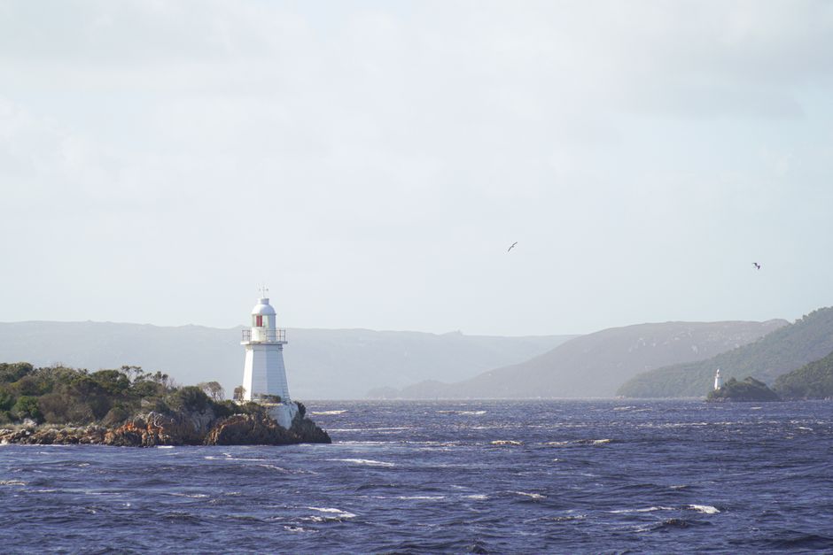 Distant view of lighthouses in Strahan, Tasmania against a backdrop of rugged landscapes and ocean waves.