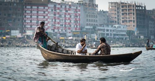 Free Three men navigate a wooden boat on a bustling river near an urban area with cityscape backdrop. Stock Photo