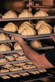 Baker placing trays of raw croissants and pastries in oven. Preparing delicious homemade bread and desserts.