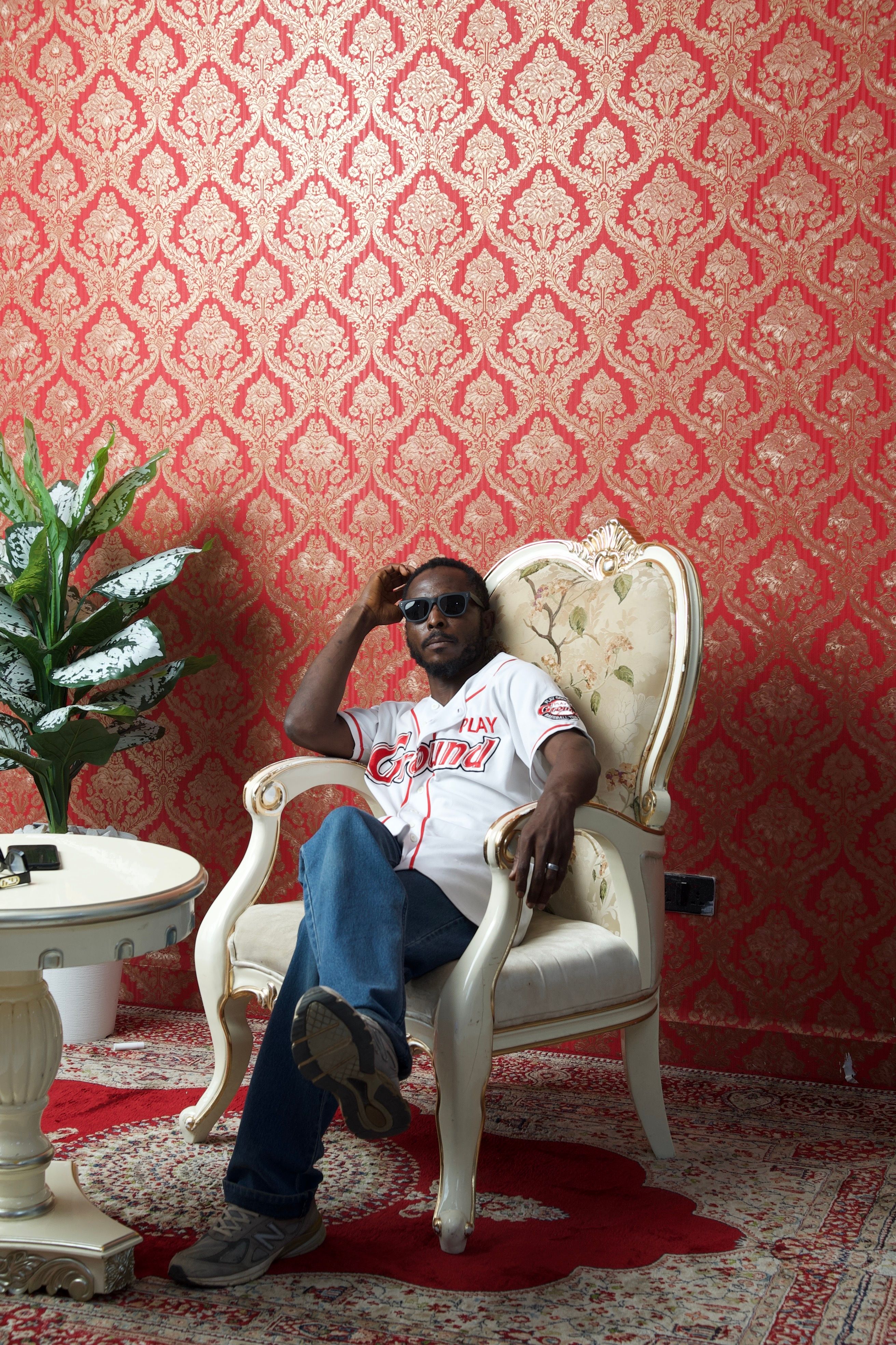 Confident man in sunglasses seated on a vintage chair in a lavishly decorated room.
