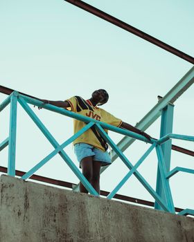 A fashionable young adult posing on a vibrant blue bridge under clear skies, showcasing urban style.
