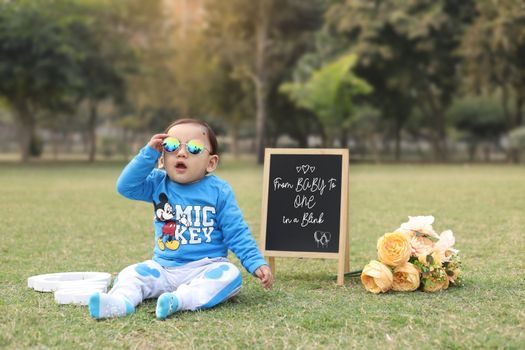 Adorable baby with sunglasses outdoors celebrating first birthday with signs and flowers.