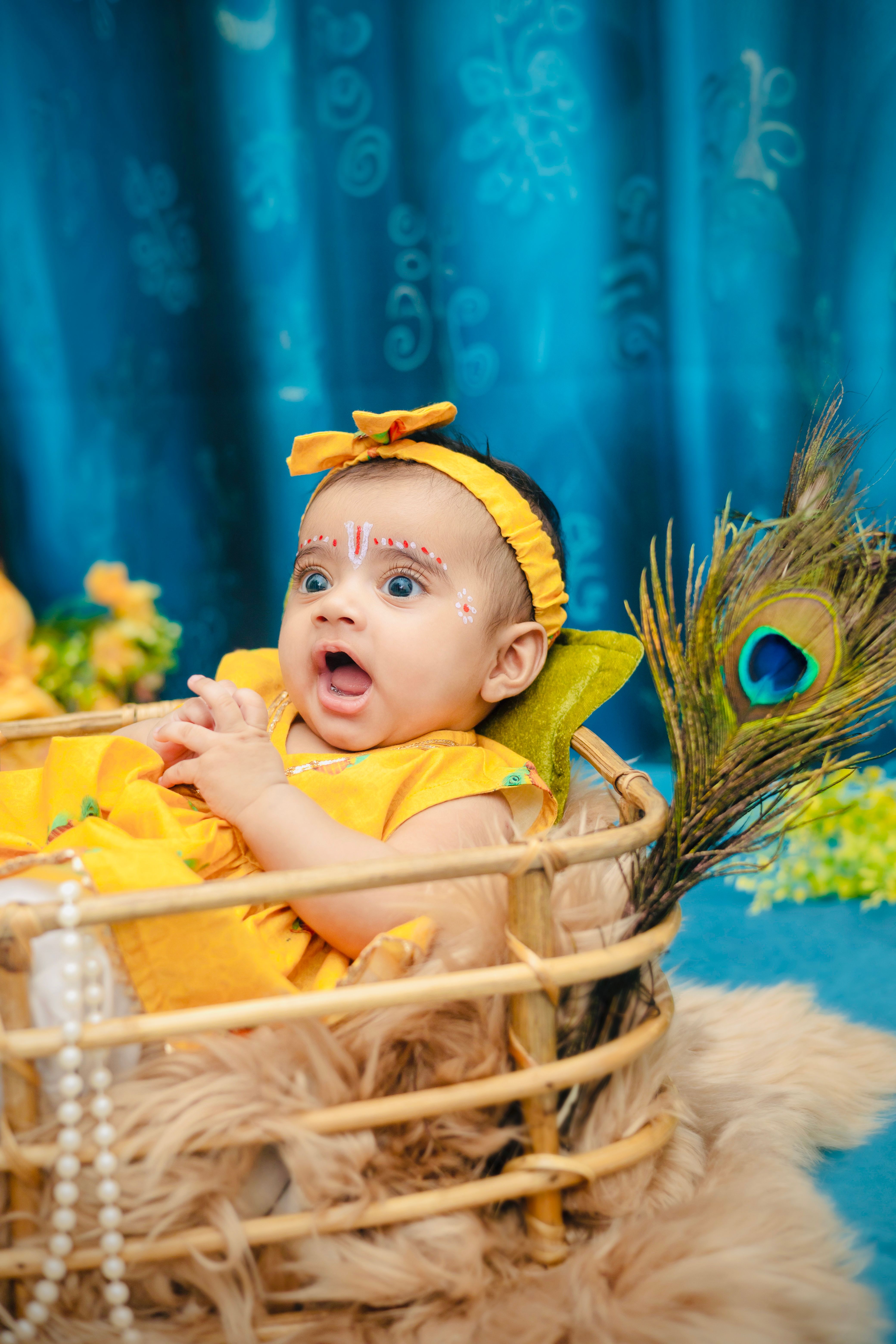 Cute baby dressed in Krishna attire with a peacock feather in a woven basket indoors.