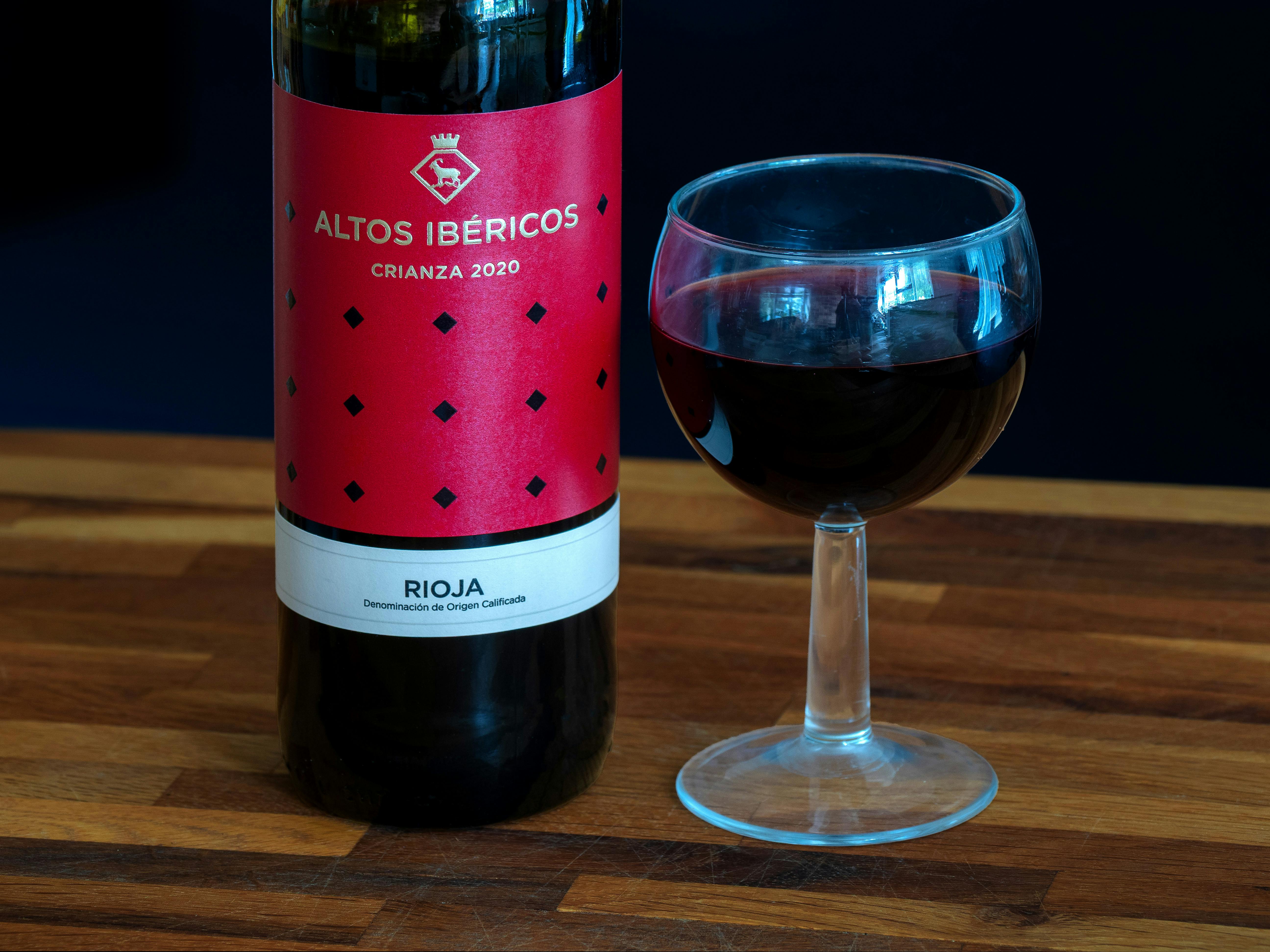Bottle of Altos Ibéricos Crianza 2020 red wine with glass on wooden table.