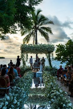 A beautiful outdoor wedding ceremony by the sea with floral decorations and palm trees at sunset.