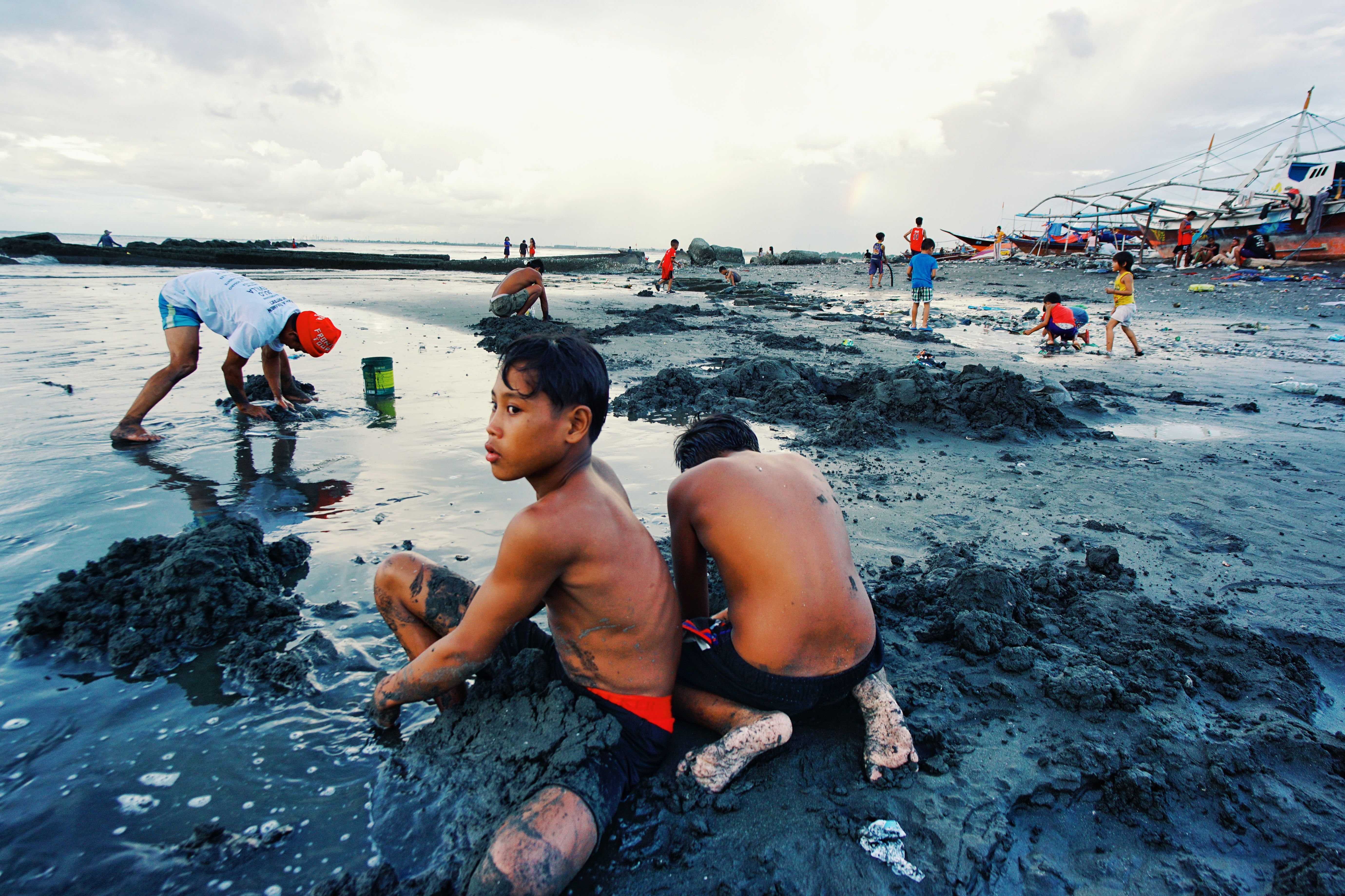 Young boys playing and digging in muddy beach area, surrounded by boats and overcast sky.