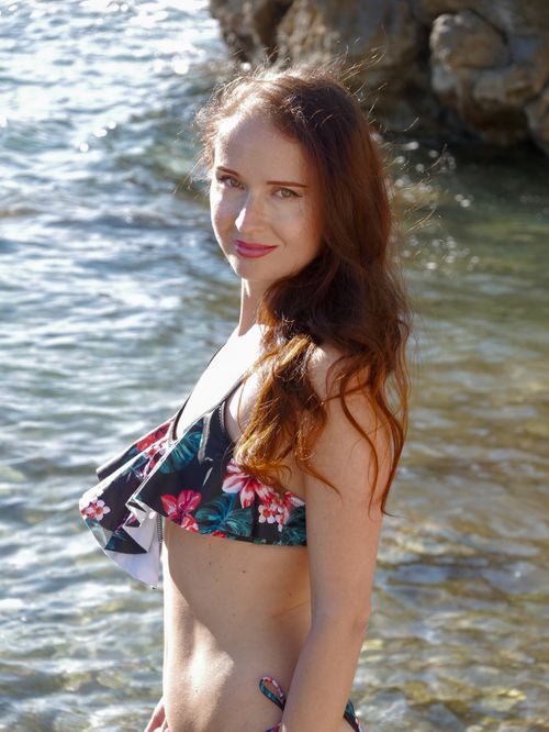 Free A woman in a floral bikini smiles by a rocky shoreline on a sunny day. Stock Photo