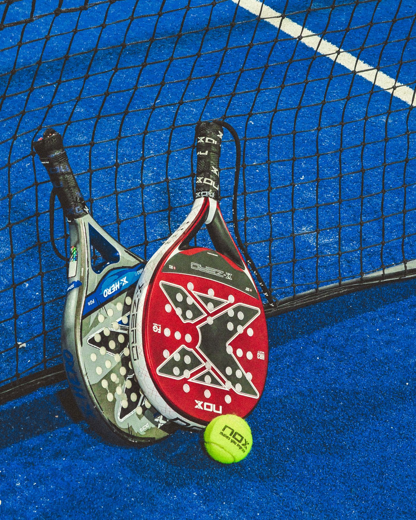 Two padel rackets and a ball resting against the net on a blue court