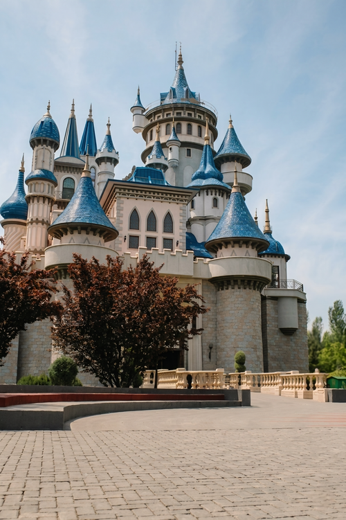 Free Elegant castle with blue spires under a clear sky, surrounded by trees and paving stones. Stock Photo