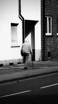 Black and white photo of an elderly man with a cane walking along a quiet urban street.